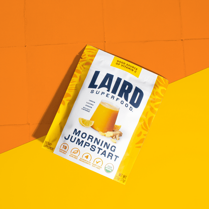 Laird Organic Morning Jumpstart powder in a 77g jar, featuring a scoop and spoon beside it, on a wooden surface with foliage.