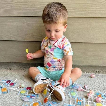 Toddler surrounded by Zollipops® Sugar Free Pineapple Lollipops in a 3.1oz package, showcasing colorful lollipops on a white background.