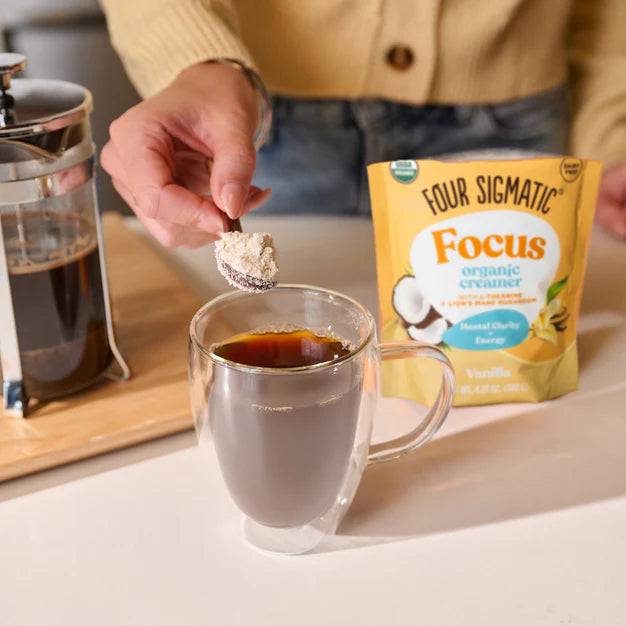 Focus Organic Vanilla Coconut Creamer from Organax Ltd, 120g, with a spoon beside a coffee cup on a wooden table.
